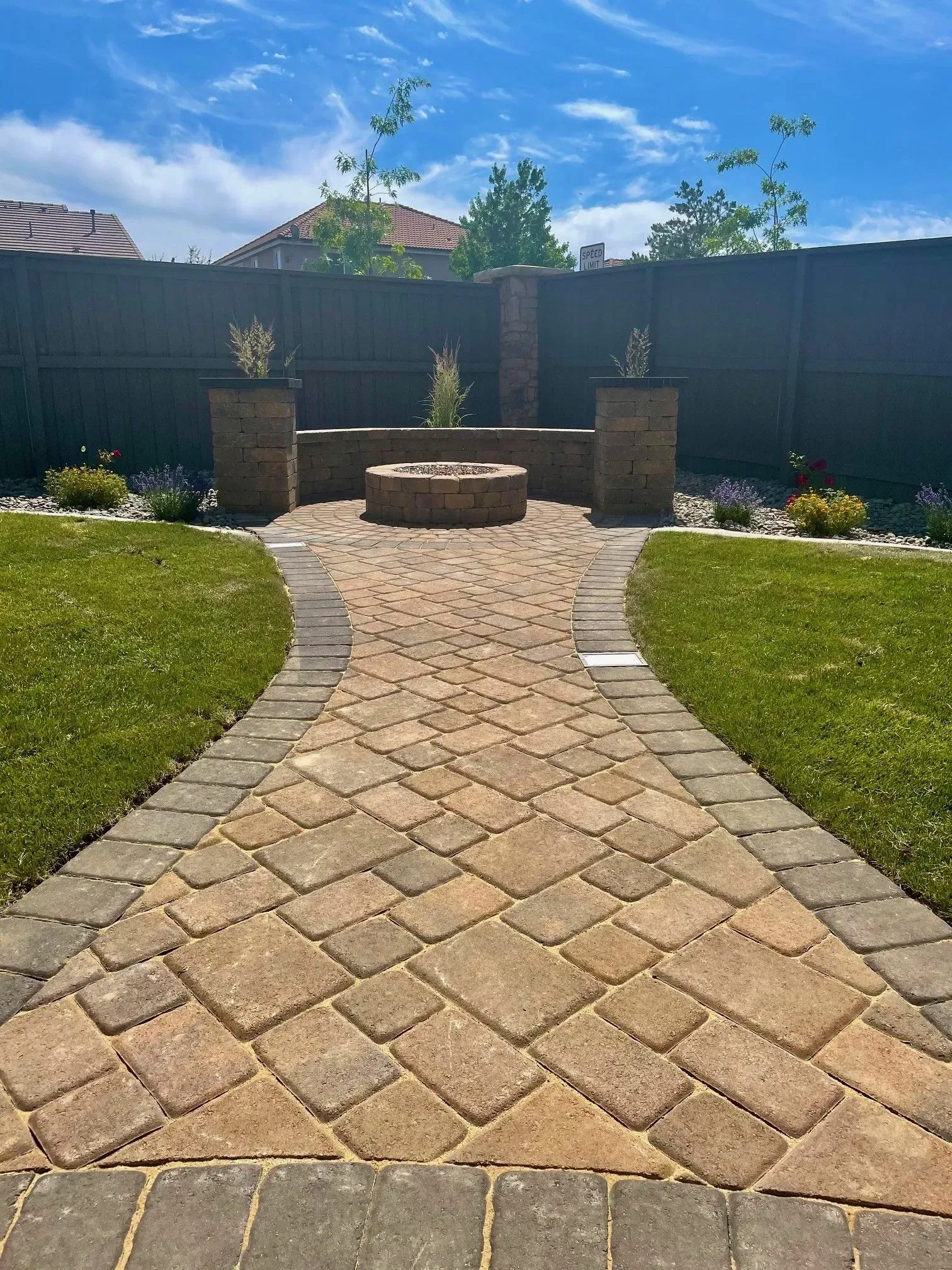 Brick patio path leading to a round fire pit and stone pillars in a sunny backyard garden