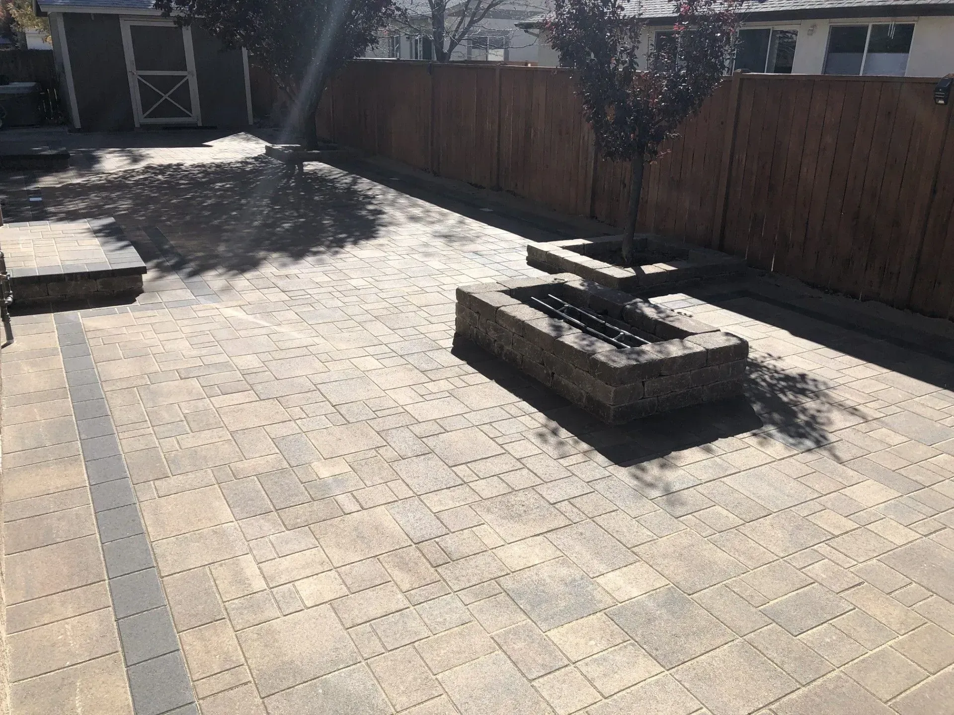 Paved backyard patio with a raised brick fire pit and wooden fence in bright sunlight