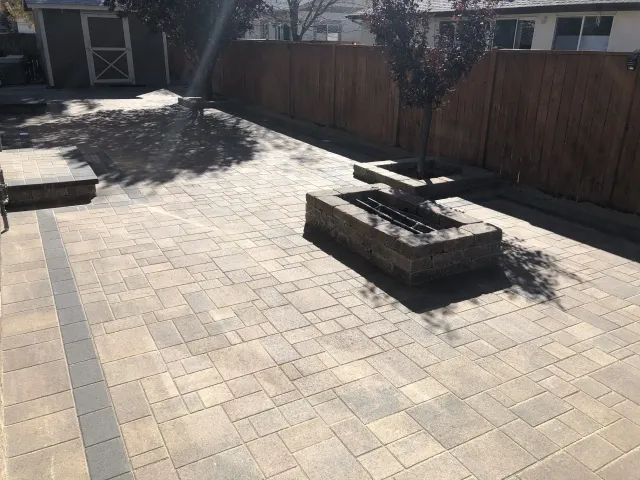 Sunny backyard patio with paver stones, raised fire pit, and wooden fence