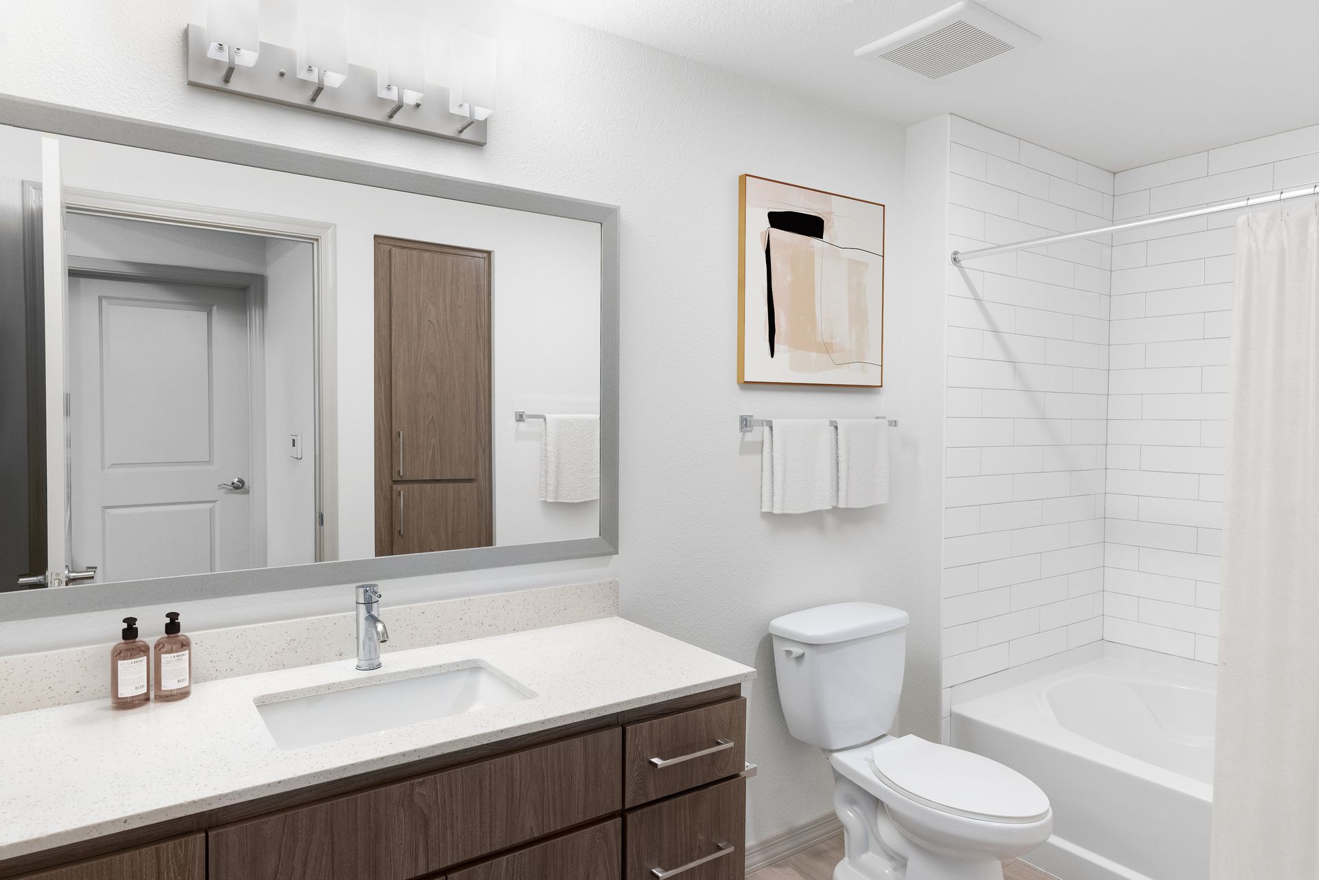 Bathroom in an apartment with a wide mirror, quartz countertop, sink, toilet, and tub/shower with white tile.