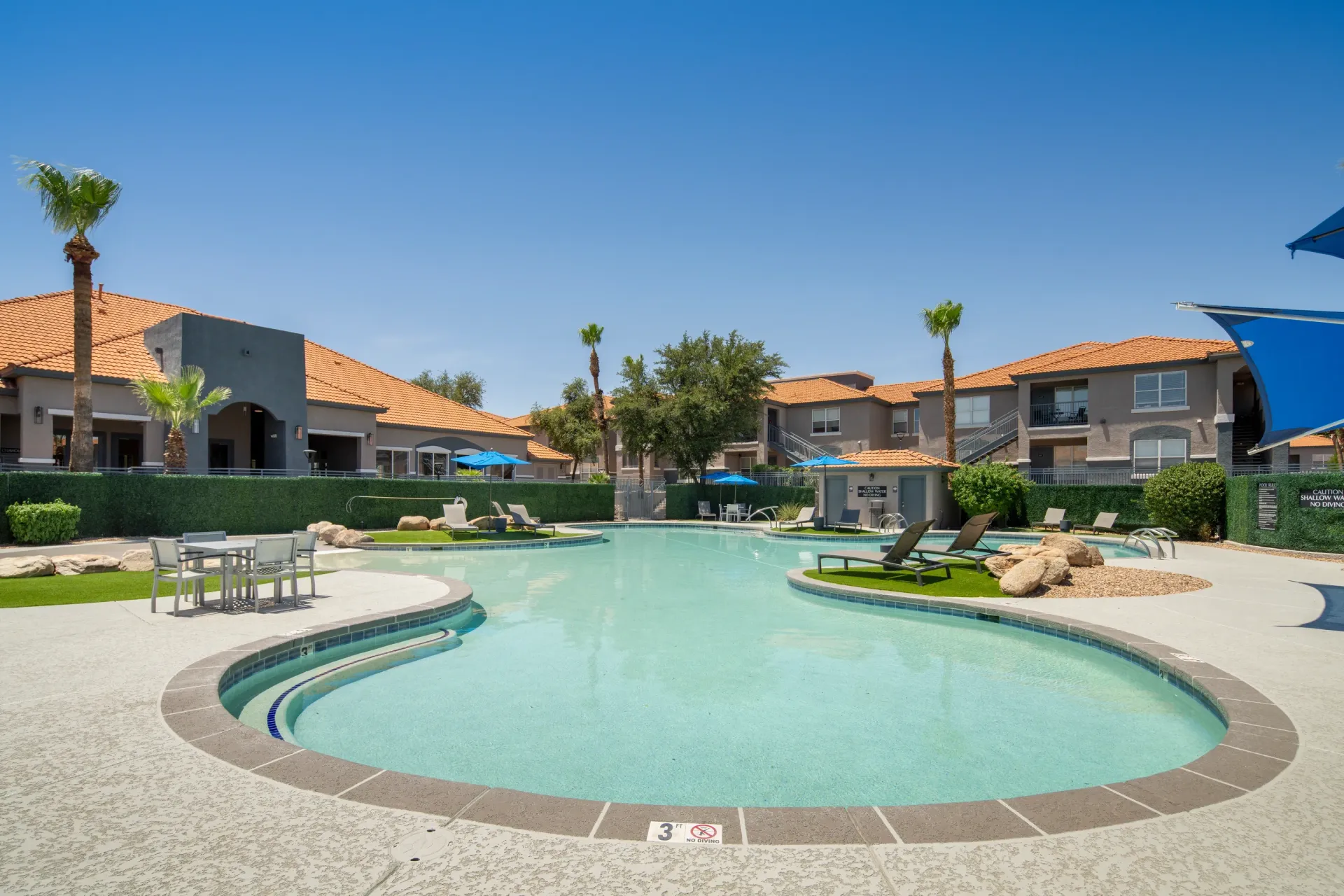 Resort-style swimming pool with lounge chairs and umbrellas, surrounded by apartment buildings and palm trees.