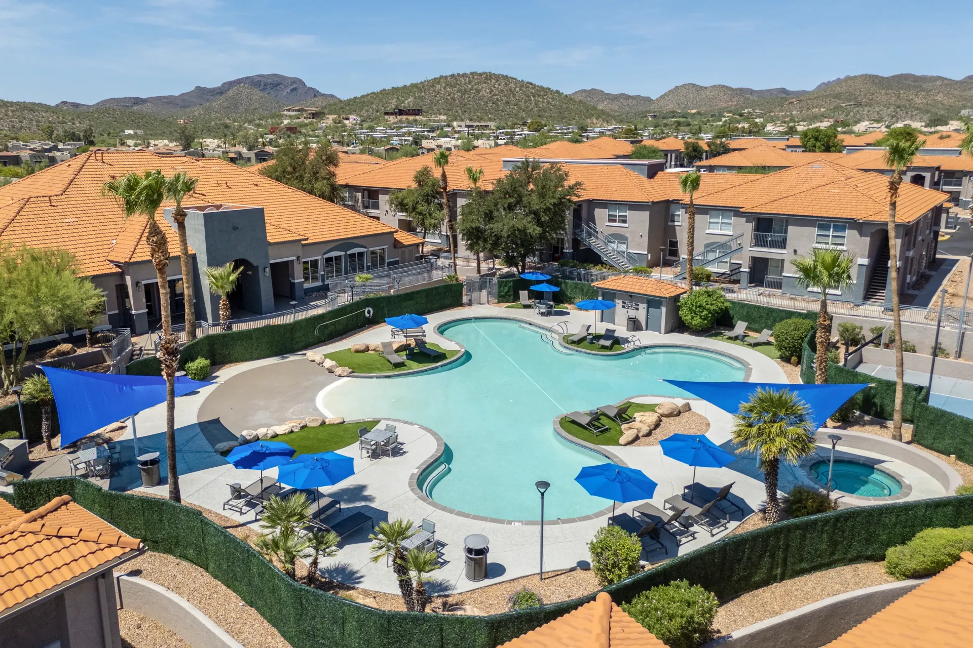 Resort-style pool and hot tub area with lounge chairs and umbrellas, surrounded by apartment buildings and landscaping at Sonoran Reserve in Tucson, AZ.