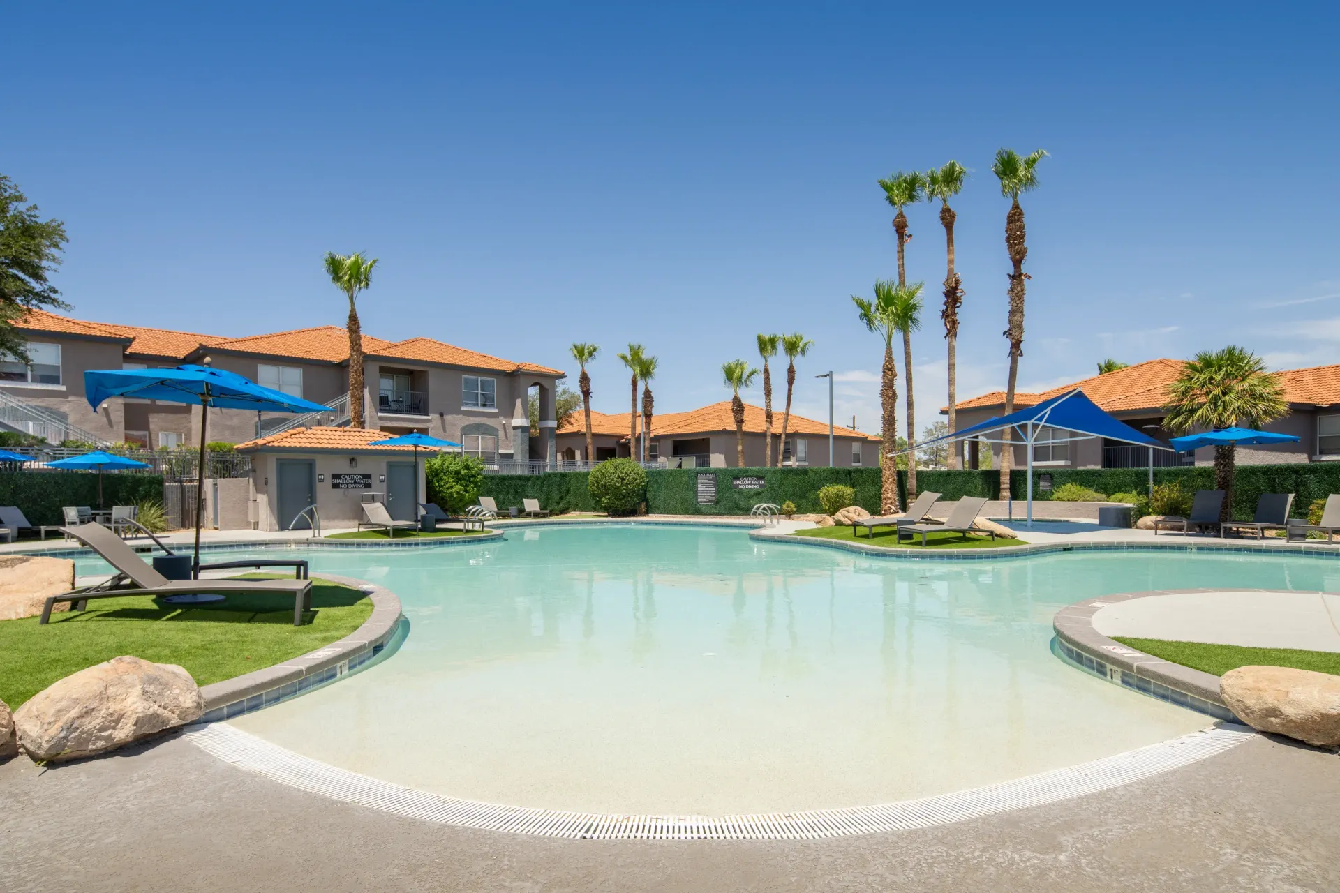 Resort-style swimming pool with lounge chairs and umbrellas, surrounded by palm trees and apartment buildings at Sonoran Reserve in Tucson, AZ.