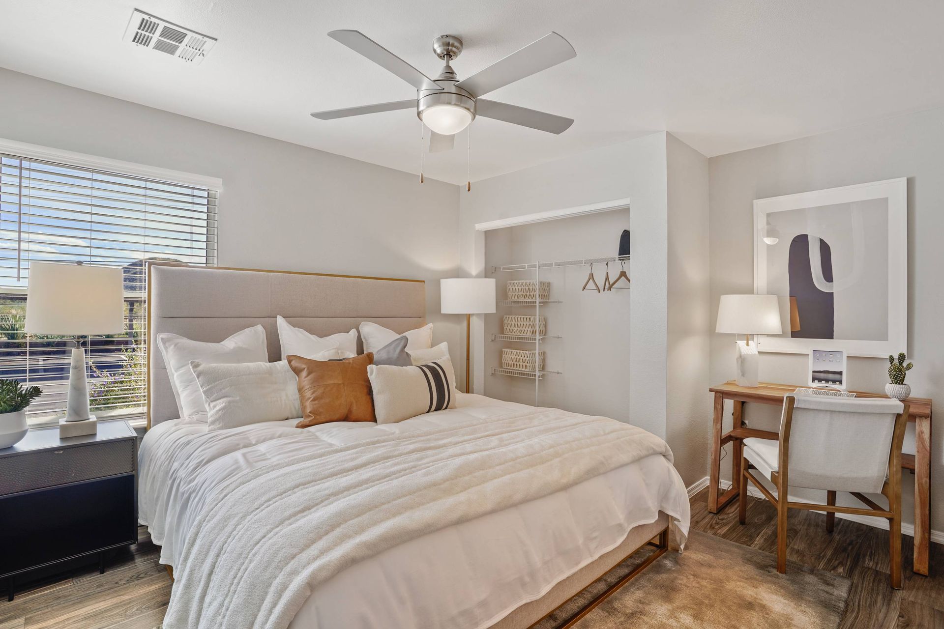 Bedroom with a bed, white bedding, nightstand, desk and chair, ceiling fan, and open closet.  See how easy it is to feel at home when you visit our welcoming pet-friendly apartments in Tucson, AZ.
