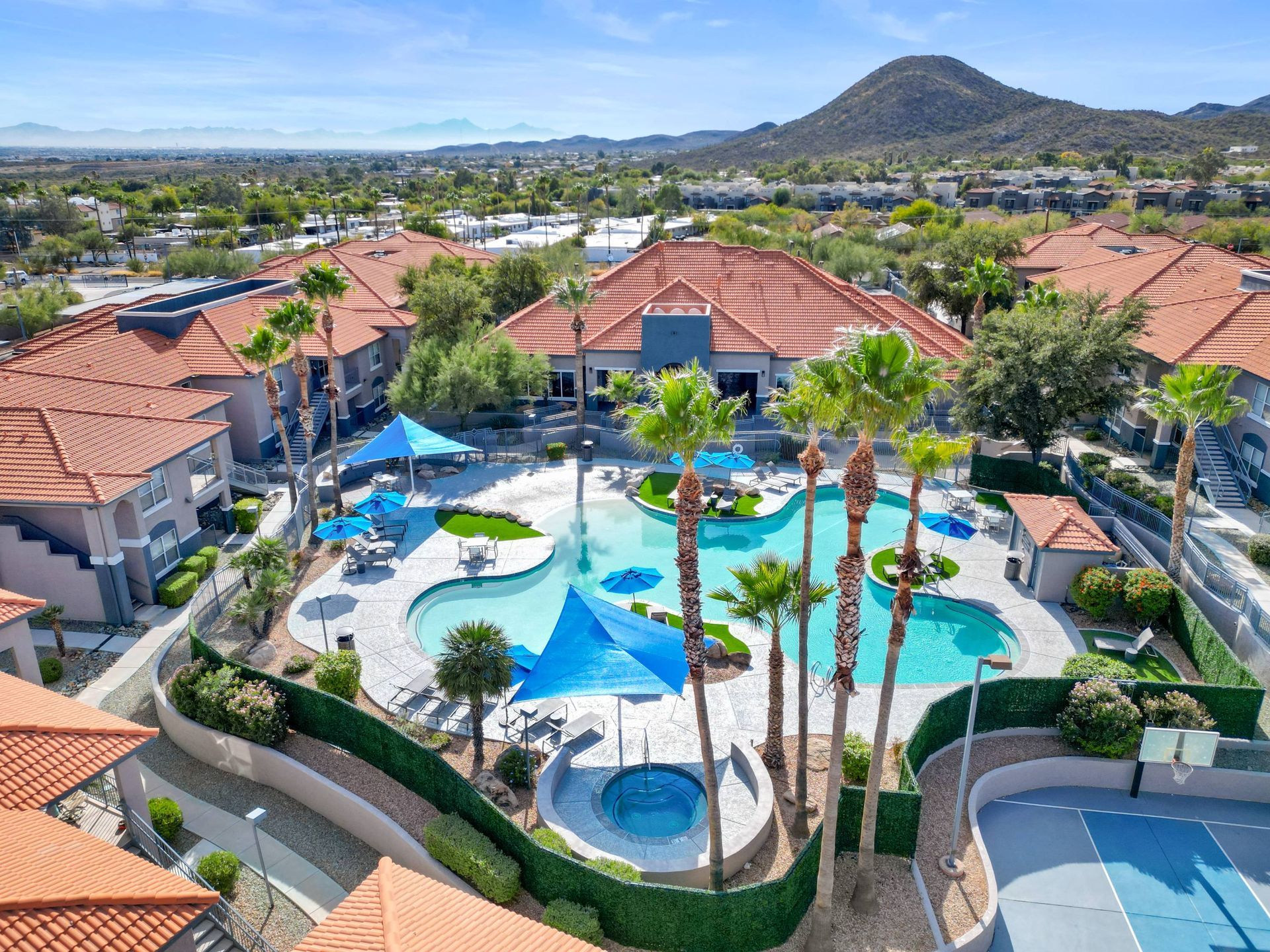 Aerial view of a resort-style apartment community pool area with palm trees and blue umbrellas.  We invite you to visit Sonoran Reserve and explore beautifully designed Tucson, AZ apartments for rent in a convenient location.
