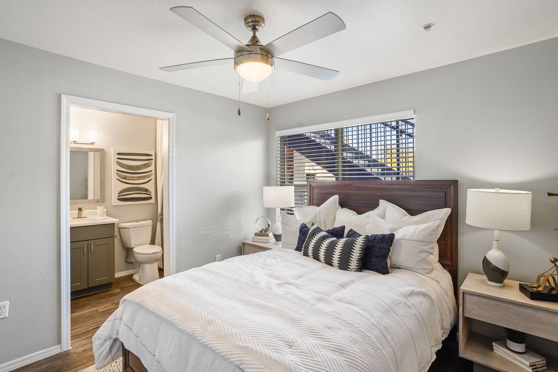 Bedroom interior with white bedding, wooden headboard, two lamps, ceiling fan, and a doorway to the bathroom.  Discover your next home when you visit Sonoran Reserve and tour our inviting Tucson, AZ, apts.