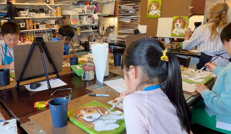 Children painting at a classroom table with art supplies and cups scattered around.
