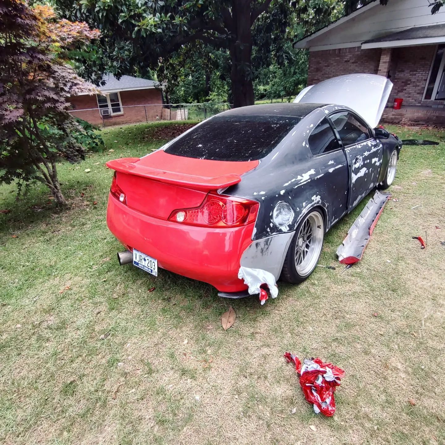 A two-door car with body panels missing, painted red, black, and silver sits on a grassy lawn with the hood open.