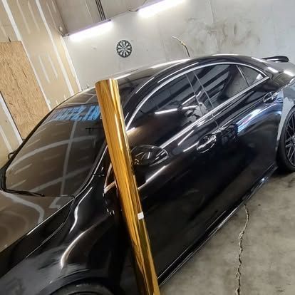 A black car with a roll of gold wrap next to it, in a garage.