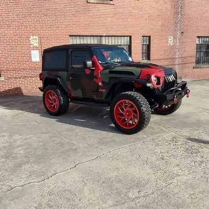 Camouflage Jeep with red accents and wheels parked outside a brick building.