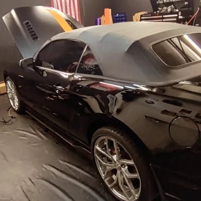 Black convertible car with gray top, hood open, and orange stripe, parked in a garage.