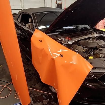 Black convertible car being wrapped in orange vinyl in a garage. Hood is open.