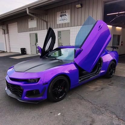 Purple sports car with doors open, parked in front of a building. Black hood and wheels.