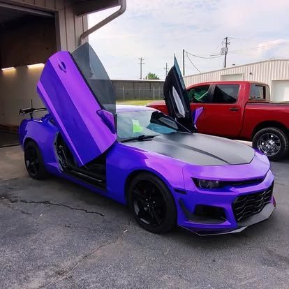Purple Chevrolet Camaro with open gullwing doors; black hood, wheels, and trim; red truck in background.
