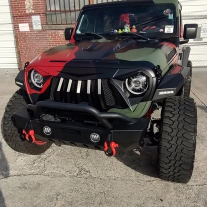 Camouflage-painted Jeep with black grill and bumper, off-road tires, and red accents parked on concrete.