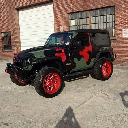 Camouflaged Jeep with red accents and rims parked in front of a brick building.
