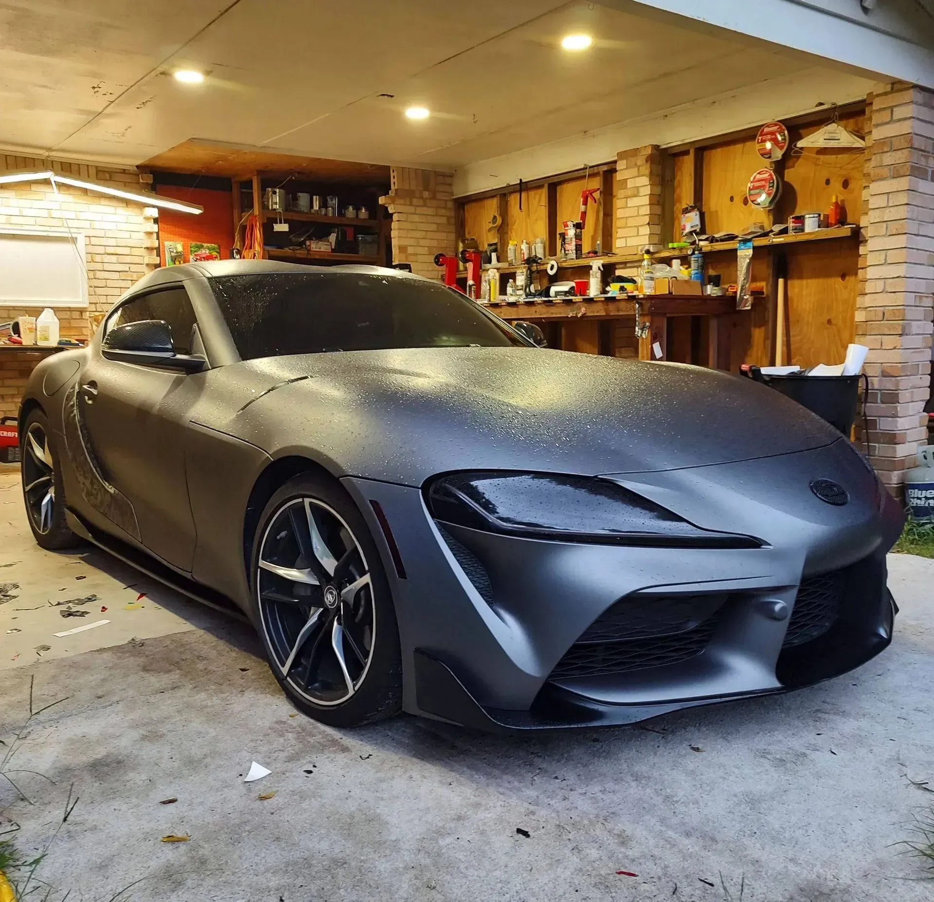 Gray Toyota GR Supra sports car parked in a garage with a workshop background.