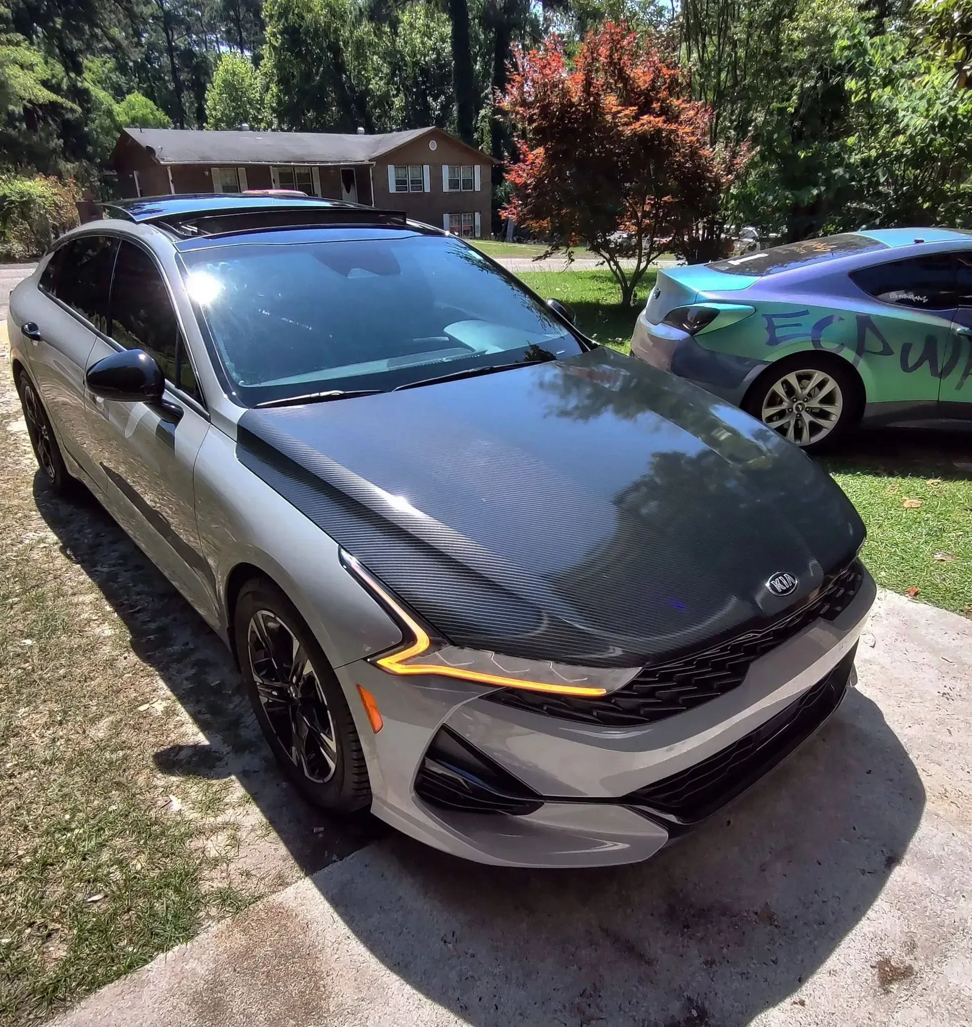 Gray Kia K5 with a carbon fiber hood, parked on a driveway with another car visible.
