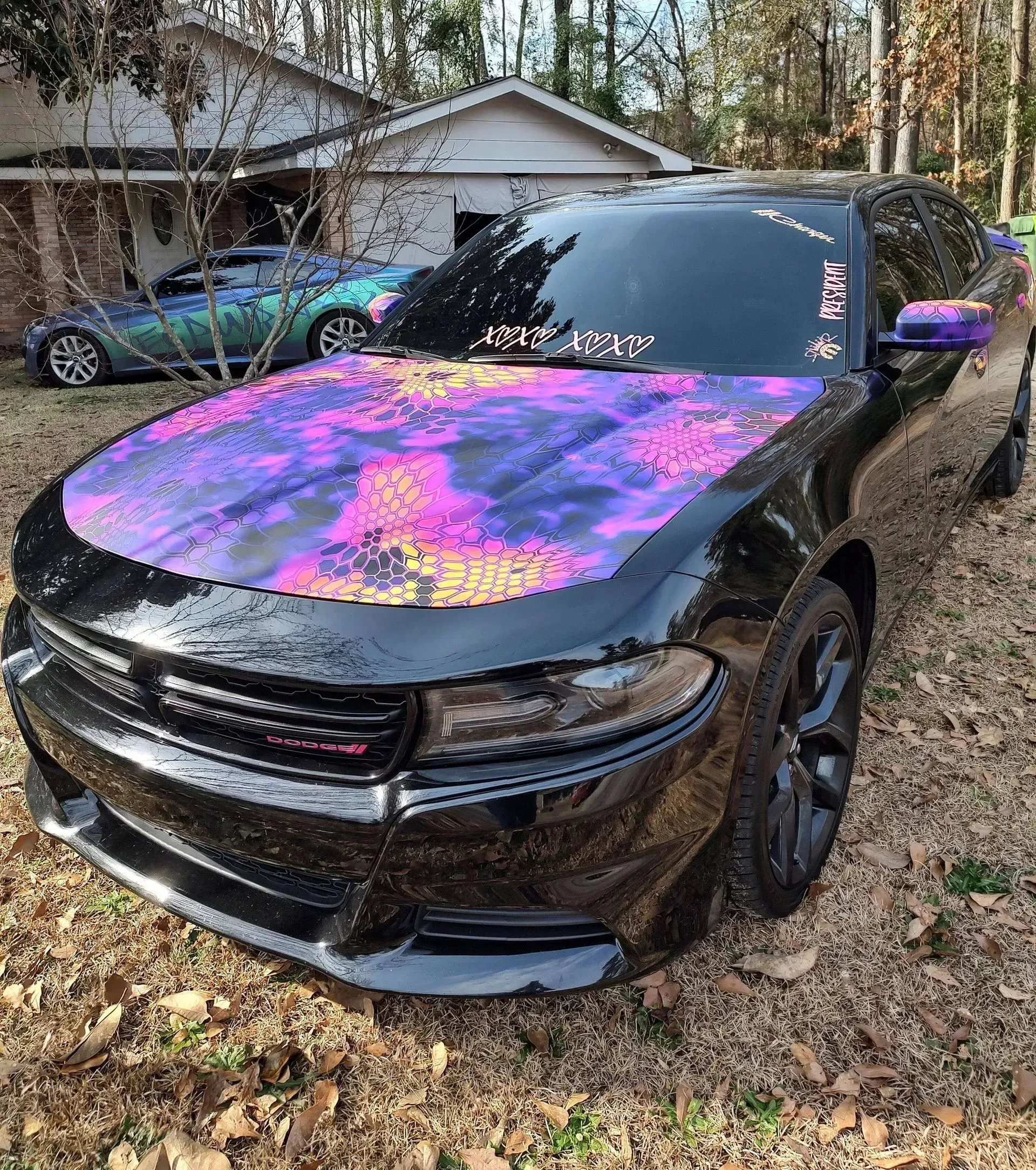 Black Dodge Charger with purple, pink, and yellow wrap on the hood. Another car is visible in the background.