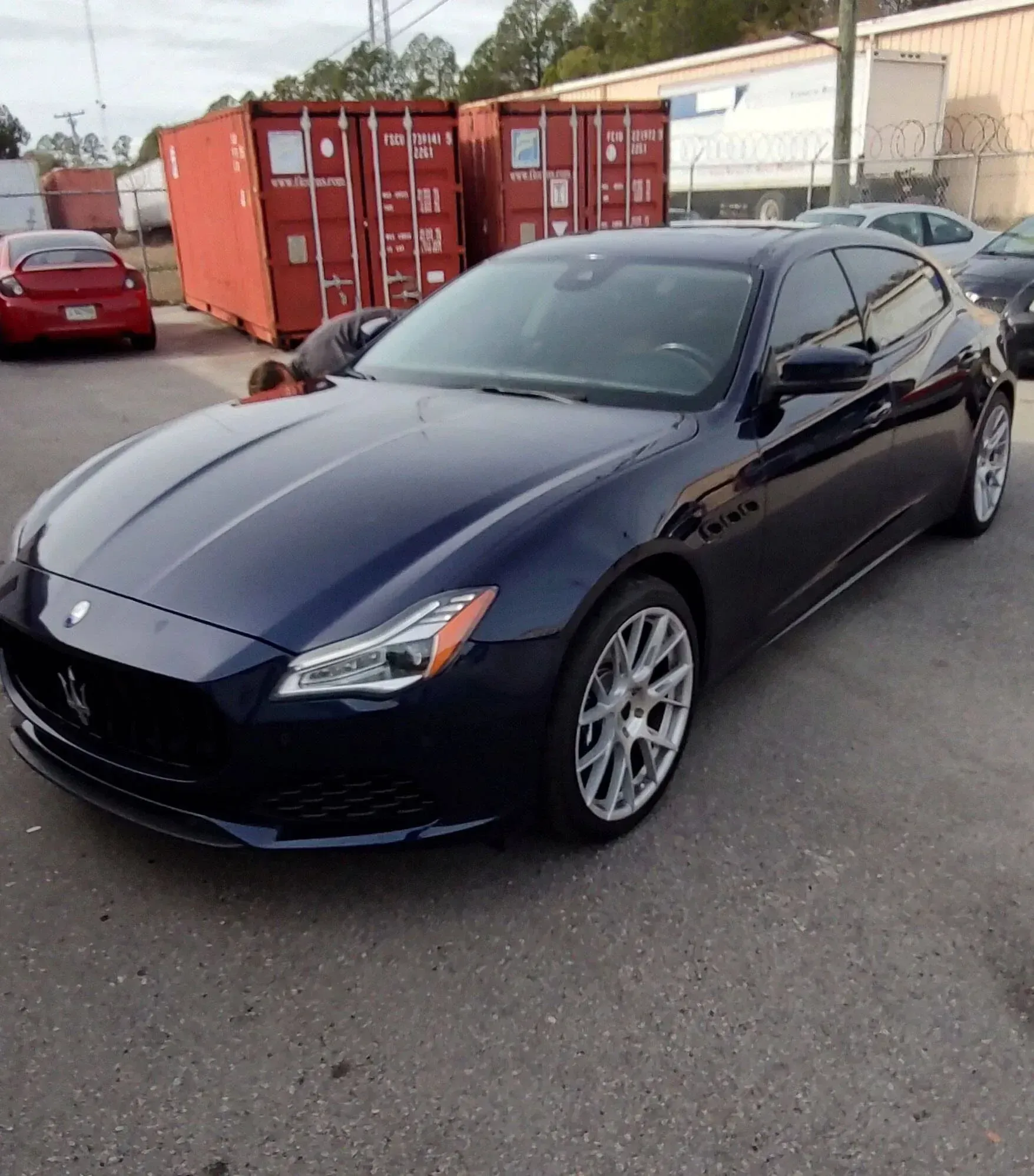 Dark blue Maserati sedan parked outdoors. Red shipping containers in the background.