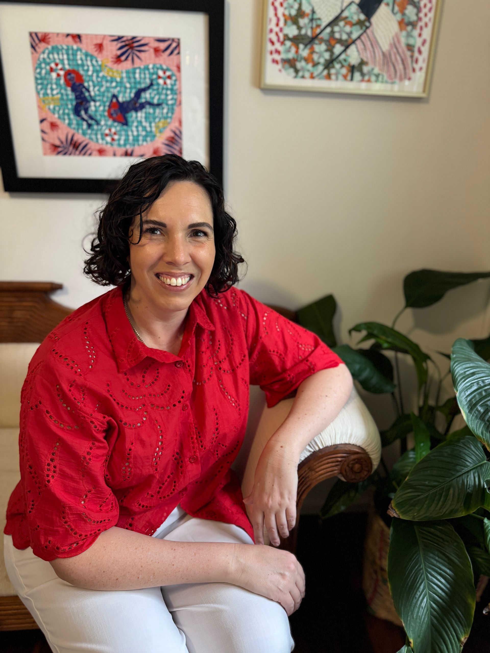 A woman in a red shirt is sitting on a couch next to a plant.