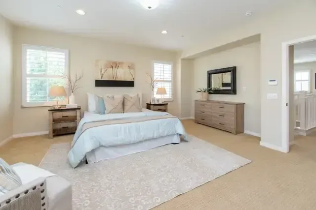Spacious beige bedroom with a large bed, dresser, mirror, and soft natural light
