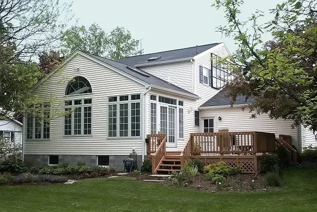 White house with large windows, wooden deck, and surrounded by trees and green lawn