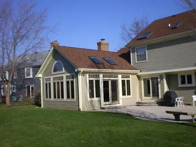 Backyard patio with a white sunroom and tan house beside a grassy lawn under a blue sky