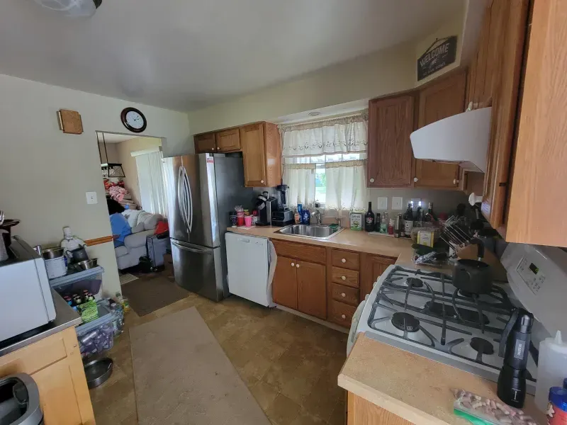 Cluttered kitchen with wooden cabinets, stainless fridge, gas stove, and a person near the back door.