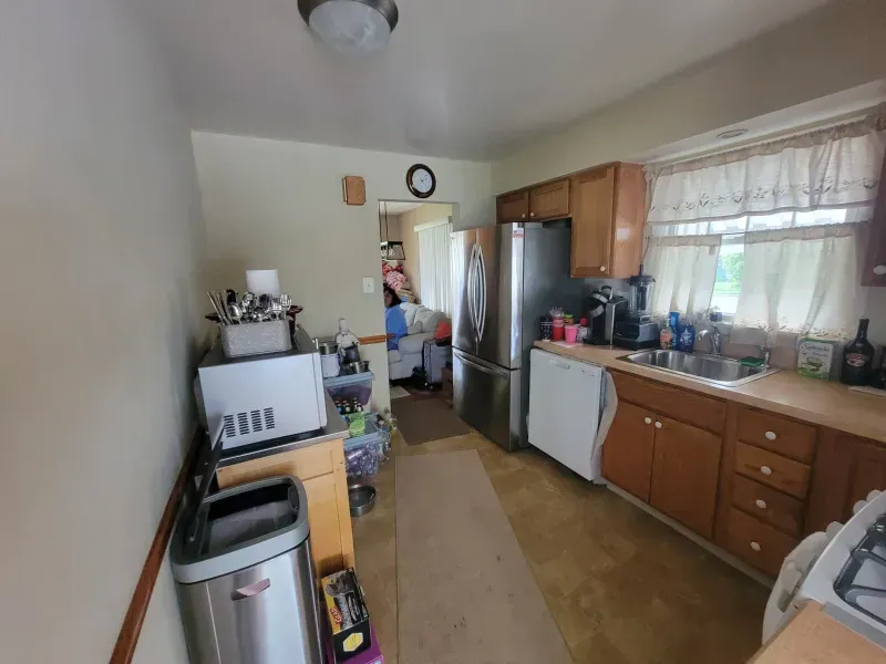Narrow kitchen with wooden cabinets, white appliances, and a person standing in the doorway.