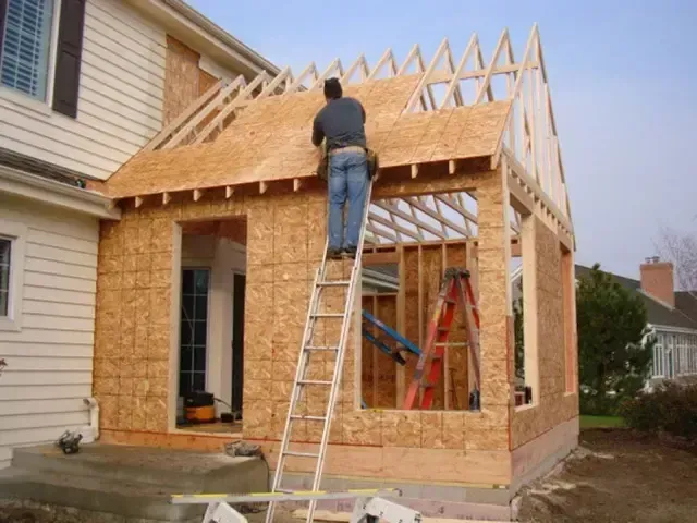 Worker building house addition with exposed wooden framing and ladder outdoors
