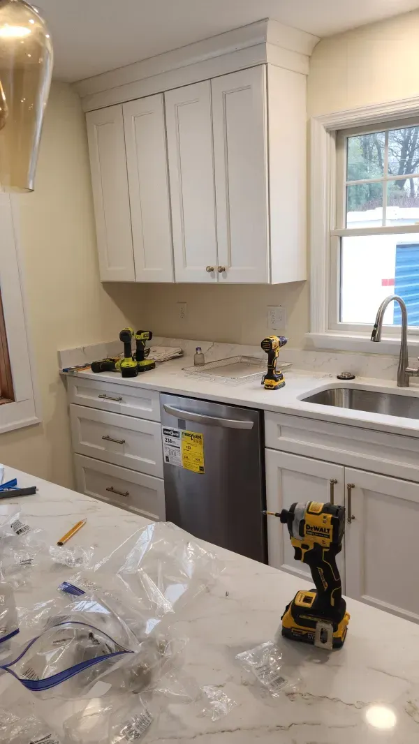 Kitchen renovation with white cabinets, marble counter, sink by window, and a yellow drill on the island.