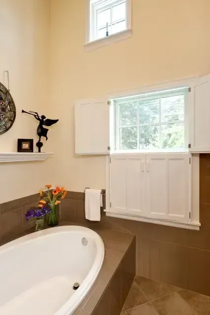 Bright bathroom with a white tub, brown tile, and a window with shutters above a small vase of flowers.