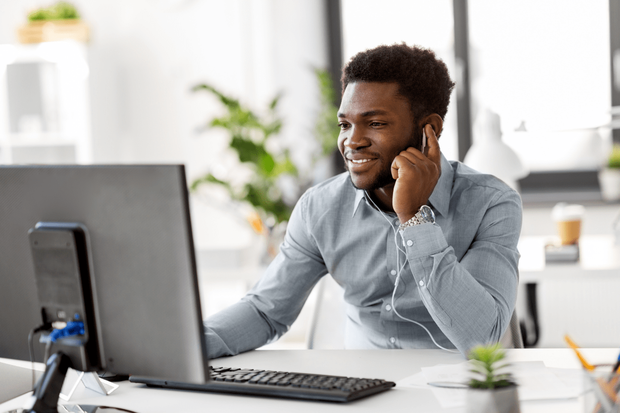 Ein Mann sitzt an einem Schreibtisch vor einem Computer und telefoniert mit seinem Mobiltelefon.