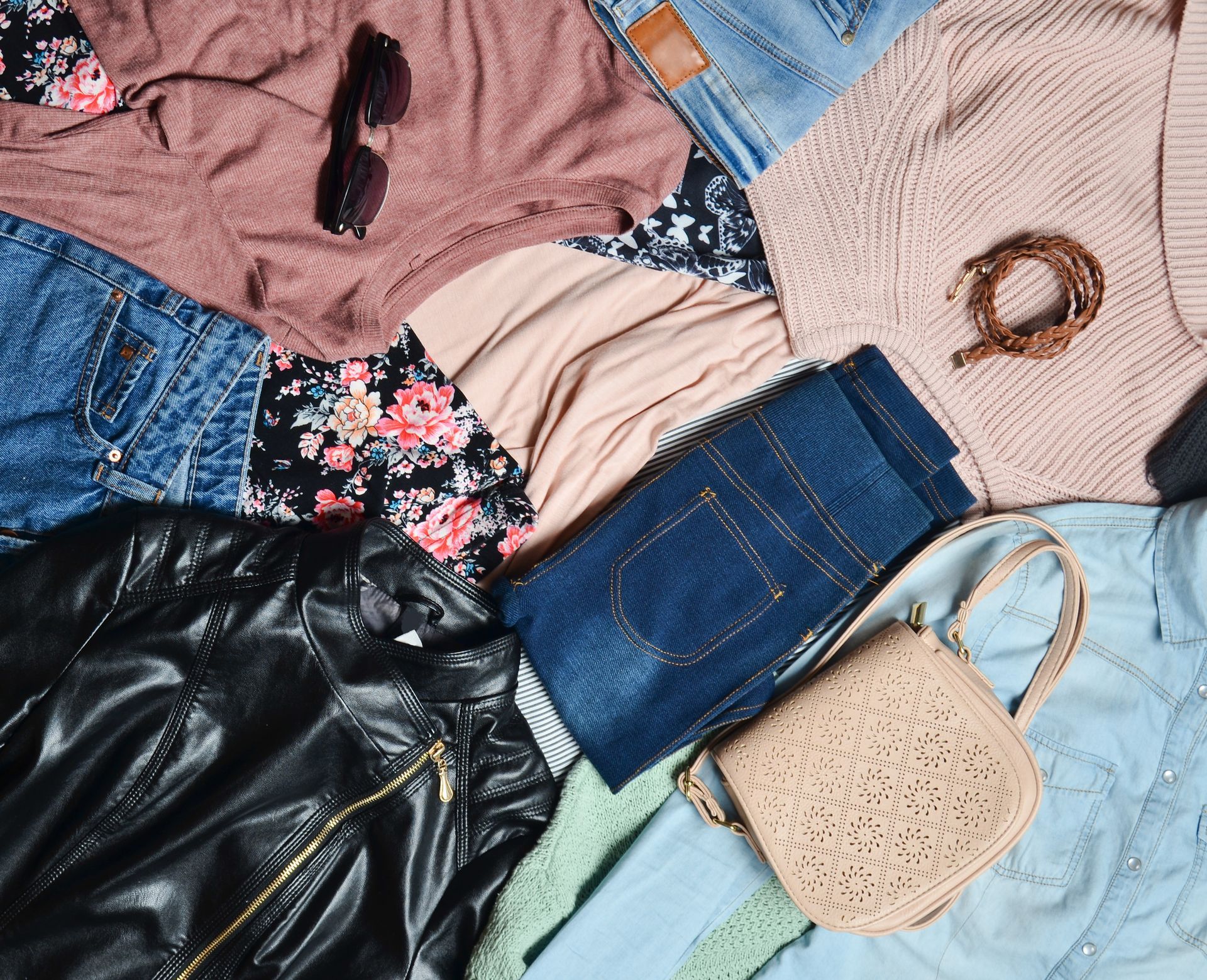 Assorted folded clothes and accessories laid out on a flat surface, including jeans, tops, jacket, sunglasses, and handbags