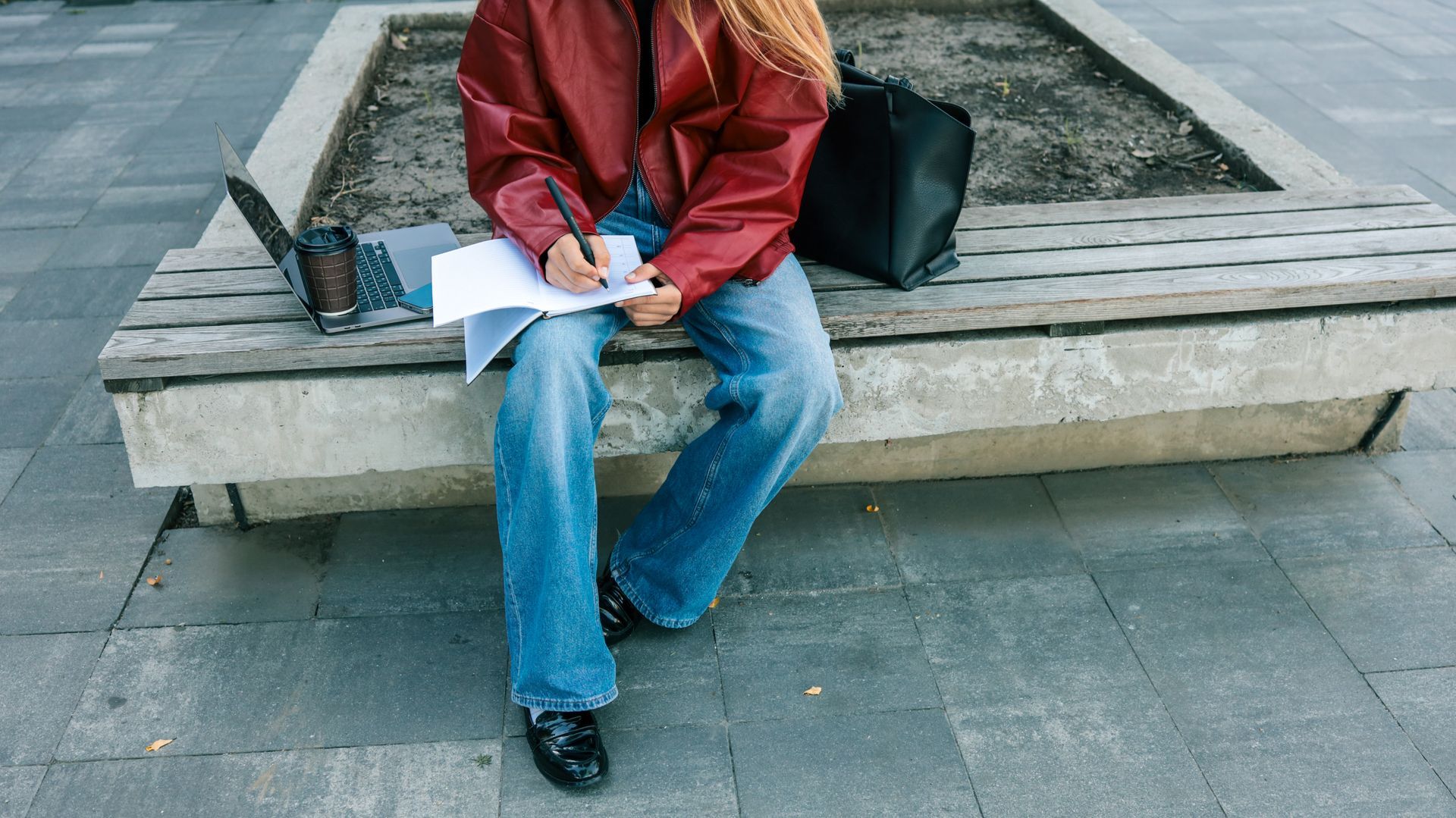 Person writing on a bench outdoors beside a backpack and water bottle.