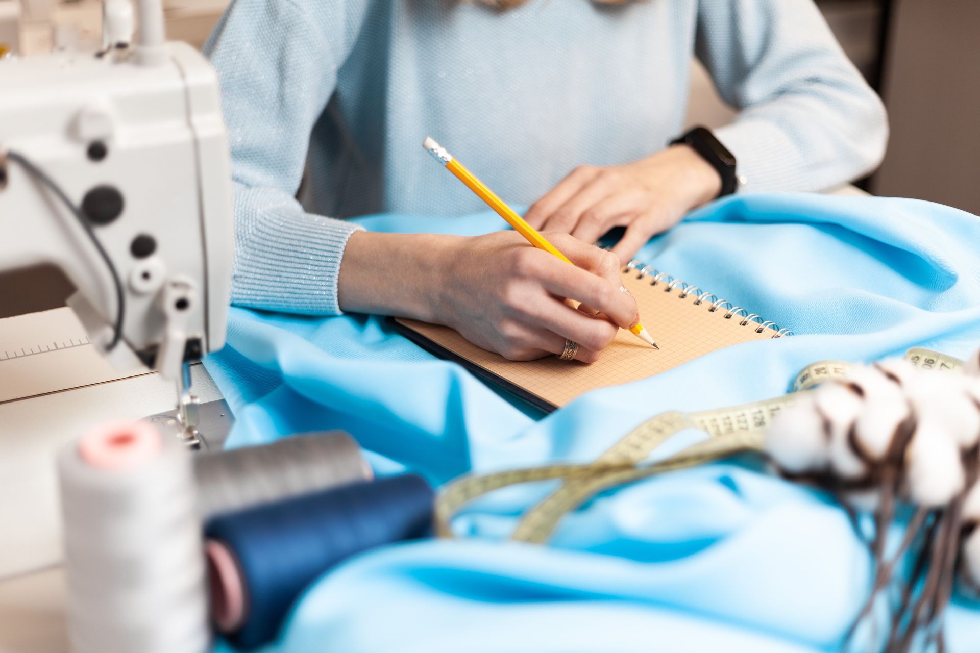 Person sketching a blue garment pattern beside a sewing machine and thread spools