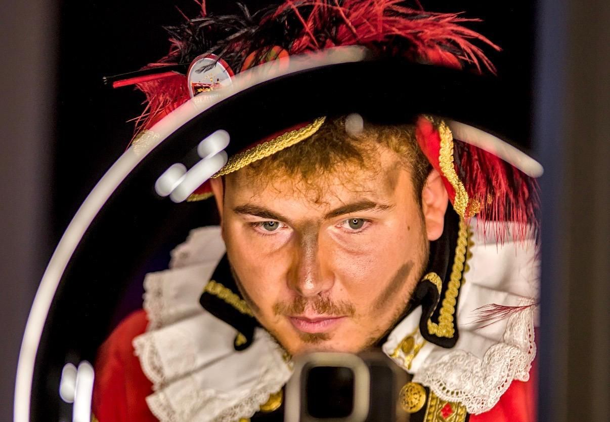 Een man in een gegolfd kostuum met rode veren, gezien door een ronde spiegel, kijkt aandachtig naar zijn spiegelbeeld.