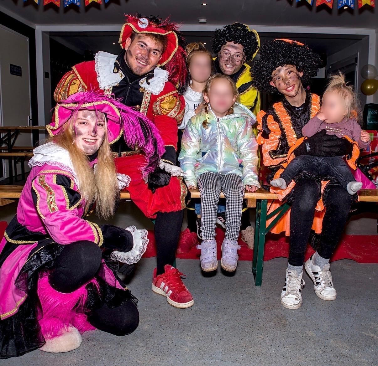 Een groep in kleurrijke kostuums poseert binnen tijdens een feestelijk evenement, met twee kinderen vooraan.