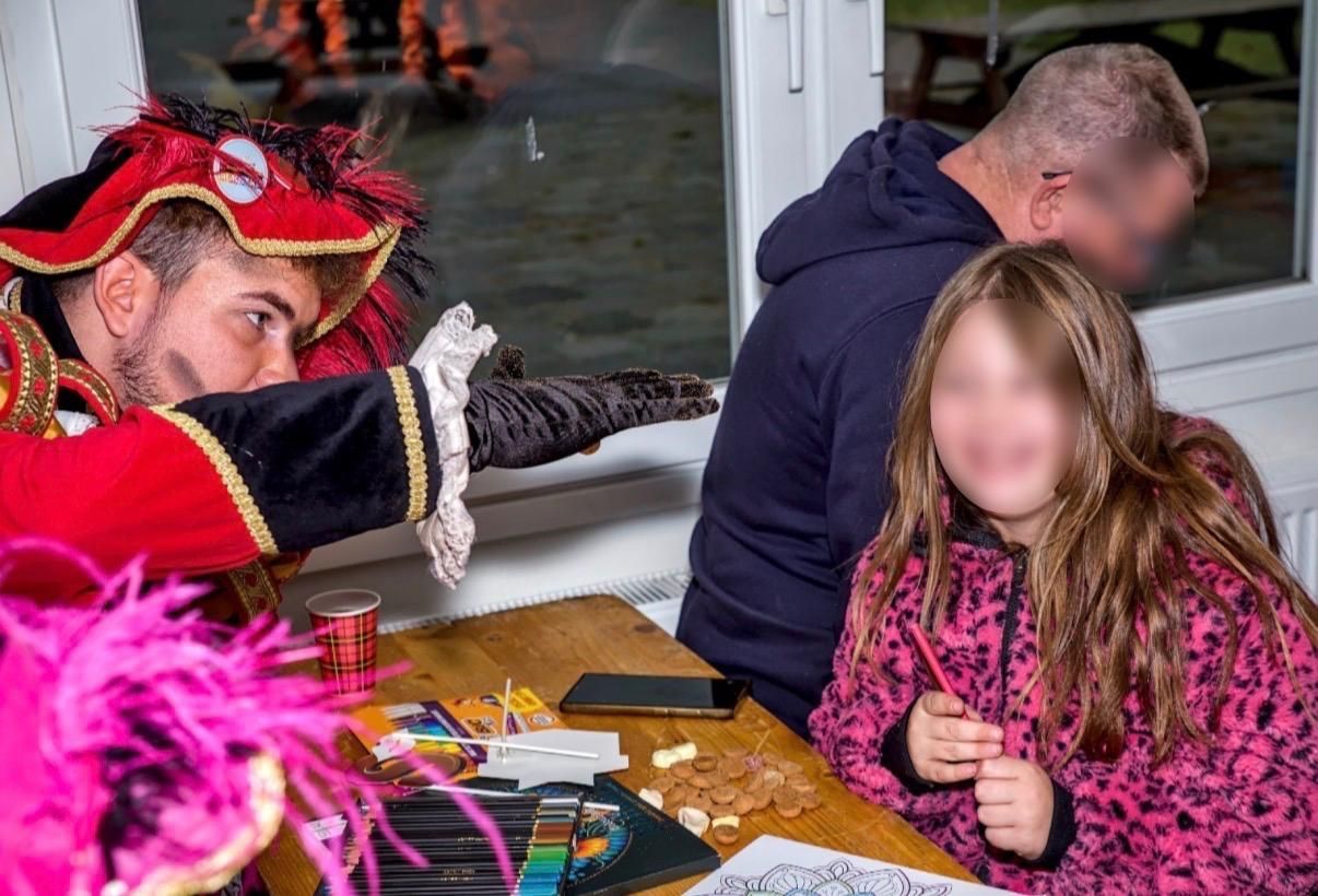 Een artiest in kostuum zit aan een tafel met een kind en een volwassene in een drukke binnenruimte.