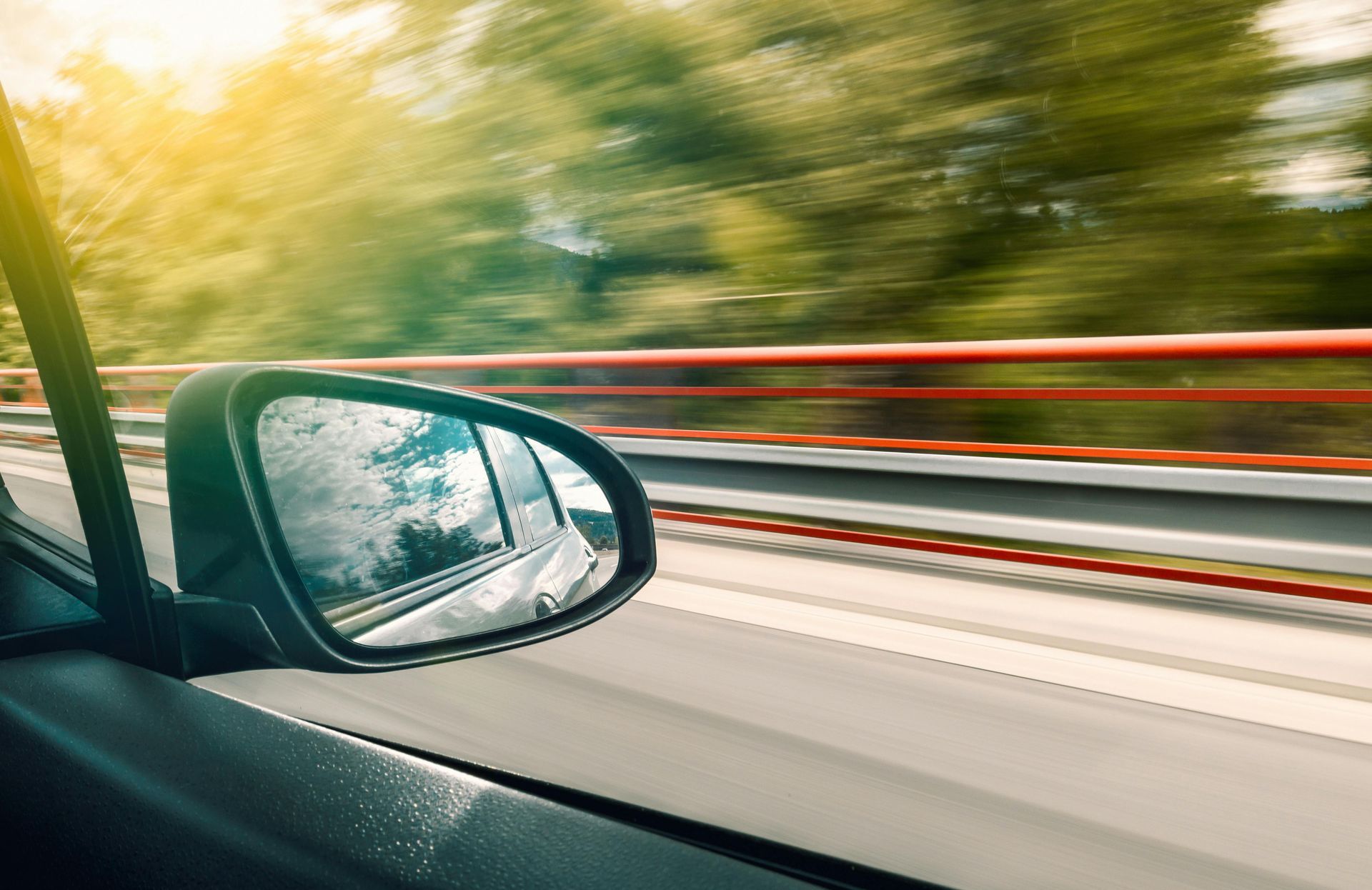 Side view mirror reflecting a blurred highway scene; motion blur, sunny day.