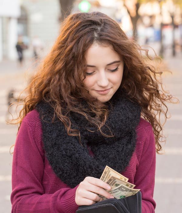 Person in a burgundy sweater and black scarf counting cash outdoors on a street.