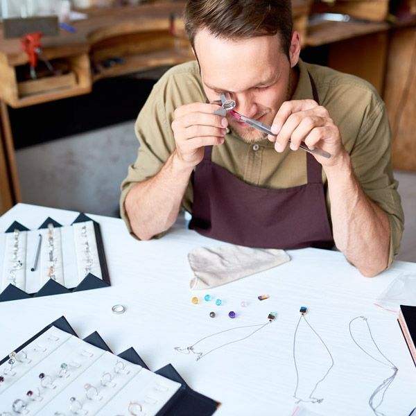 Person examining a small craft piece at a worktable with beads, tools, and sketches spread out.