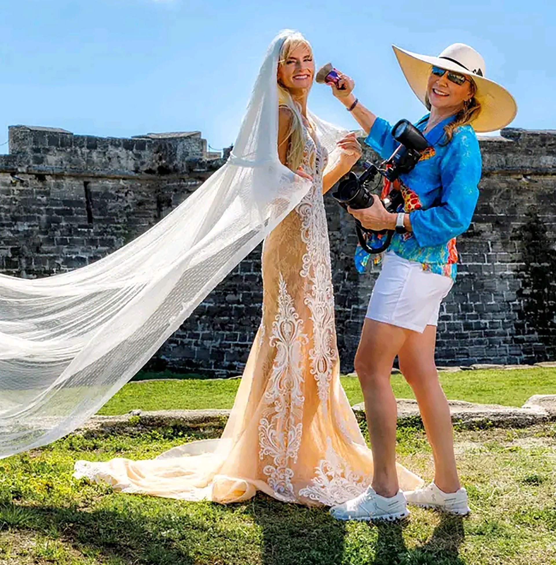Bride in a long white veil and gown posing with a photographer beside a stone wall on a sunny day
