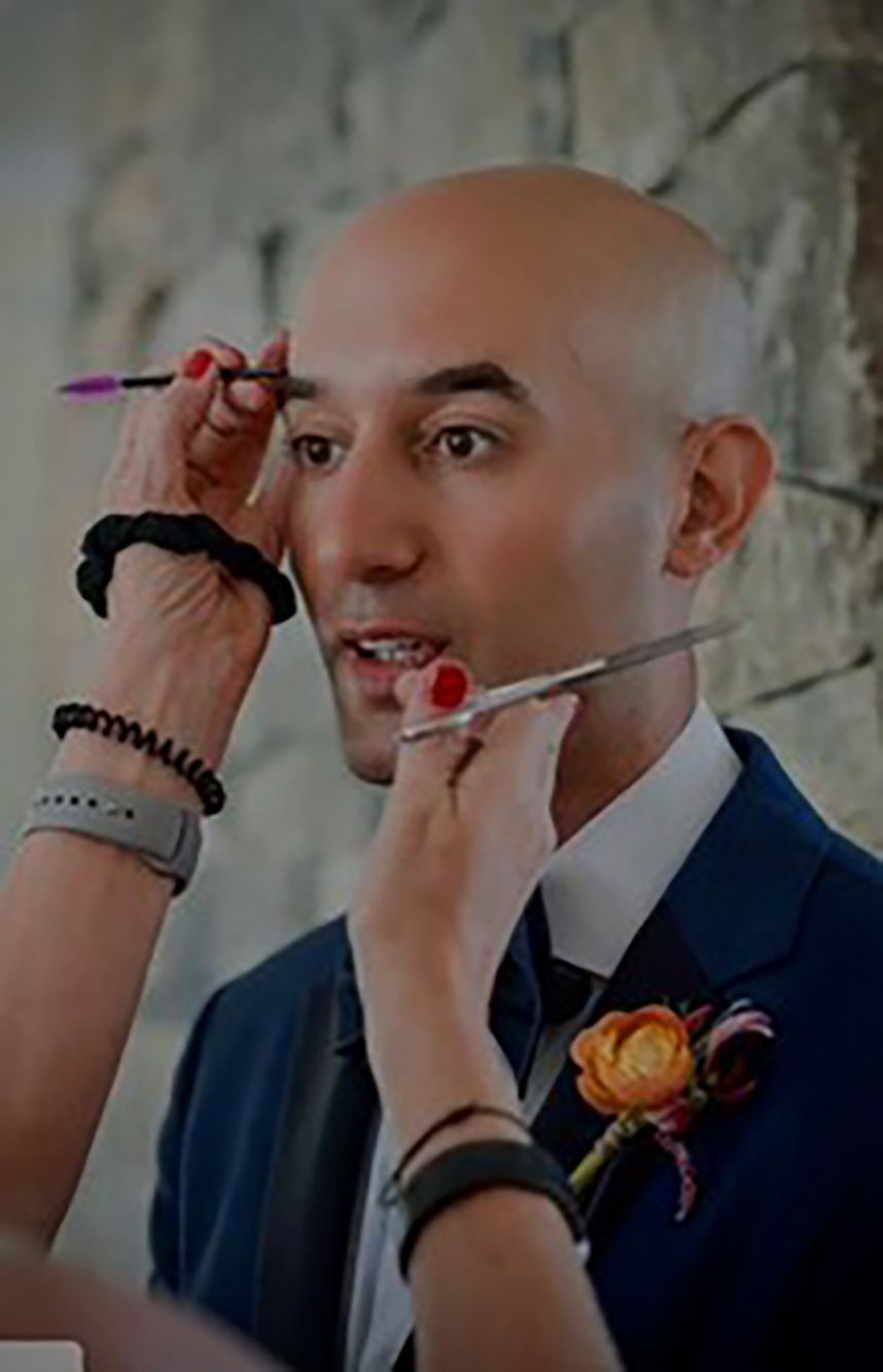 Man getting makeup applied with brushes, wearing a navy suit and boutonniere before an event