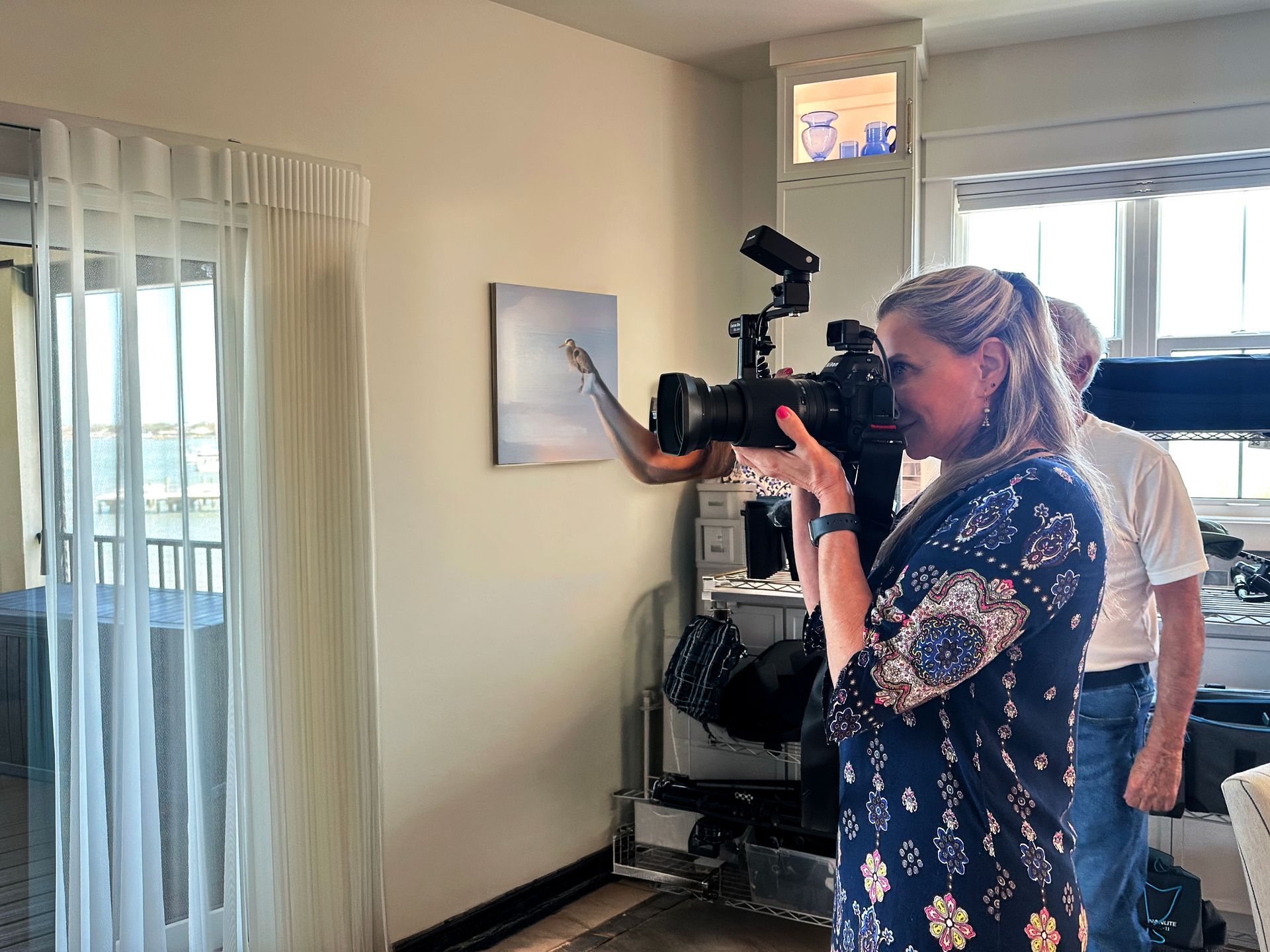 Photographer taking a photo indoors near a window with sheer curtains and a framed wall print