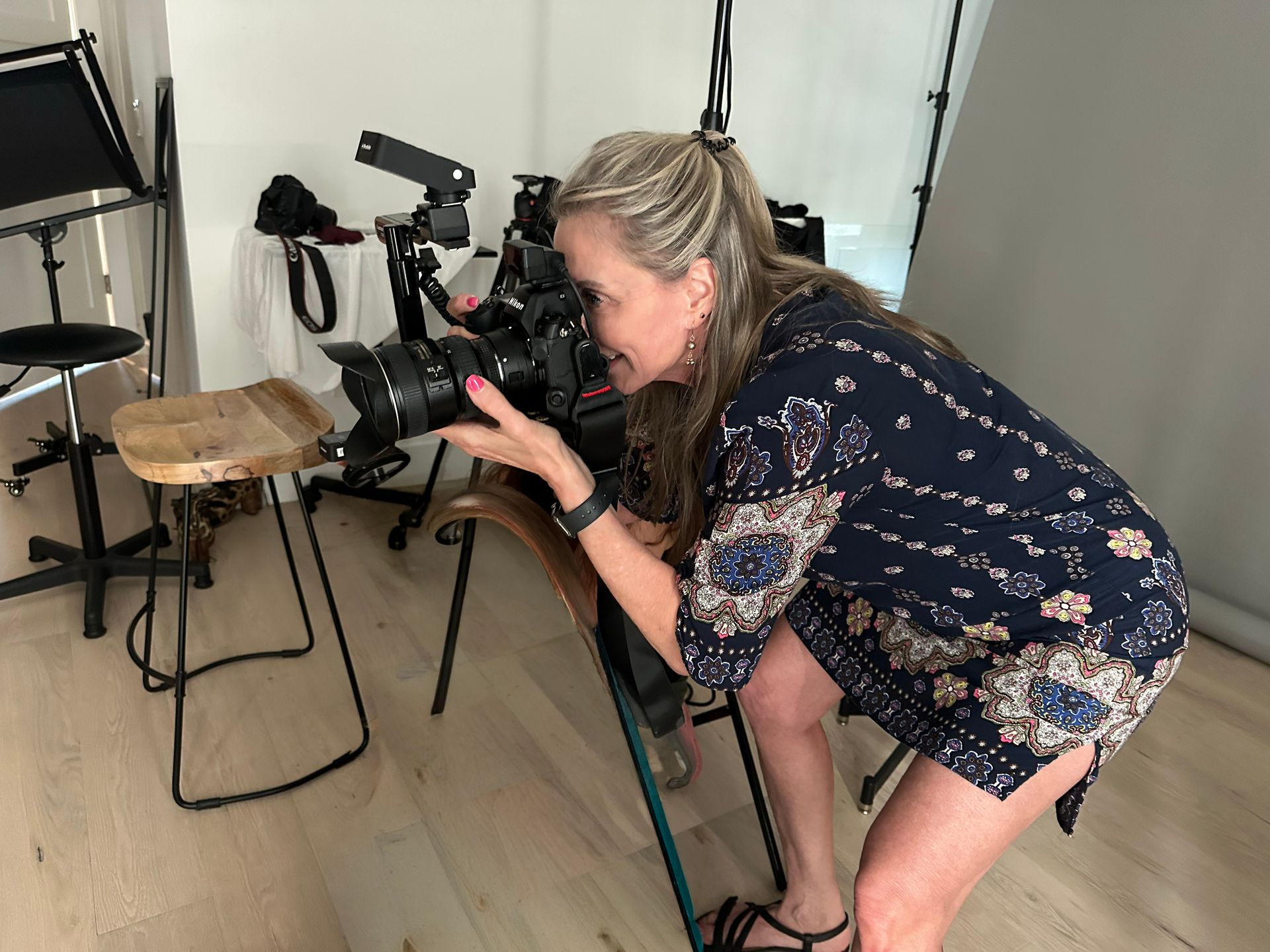 Photographer crouches beside a camera on a tripod in a bright studio, aiming at a subject off-frame