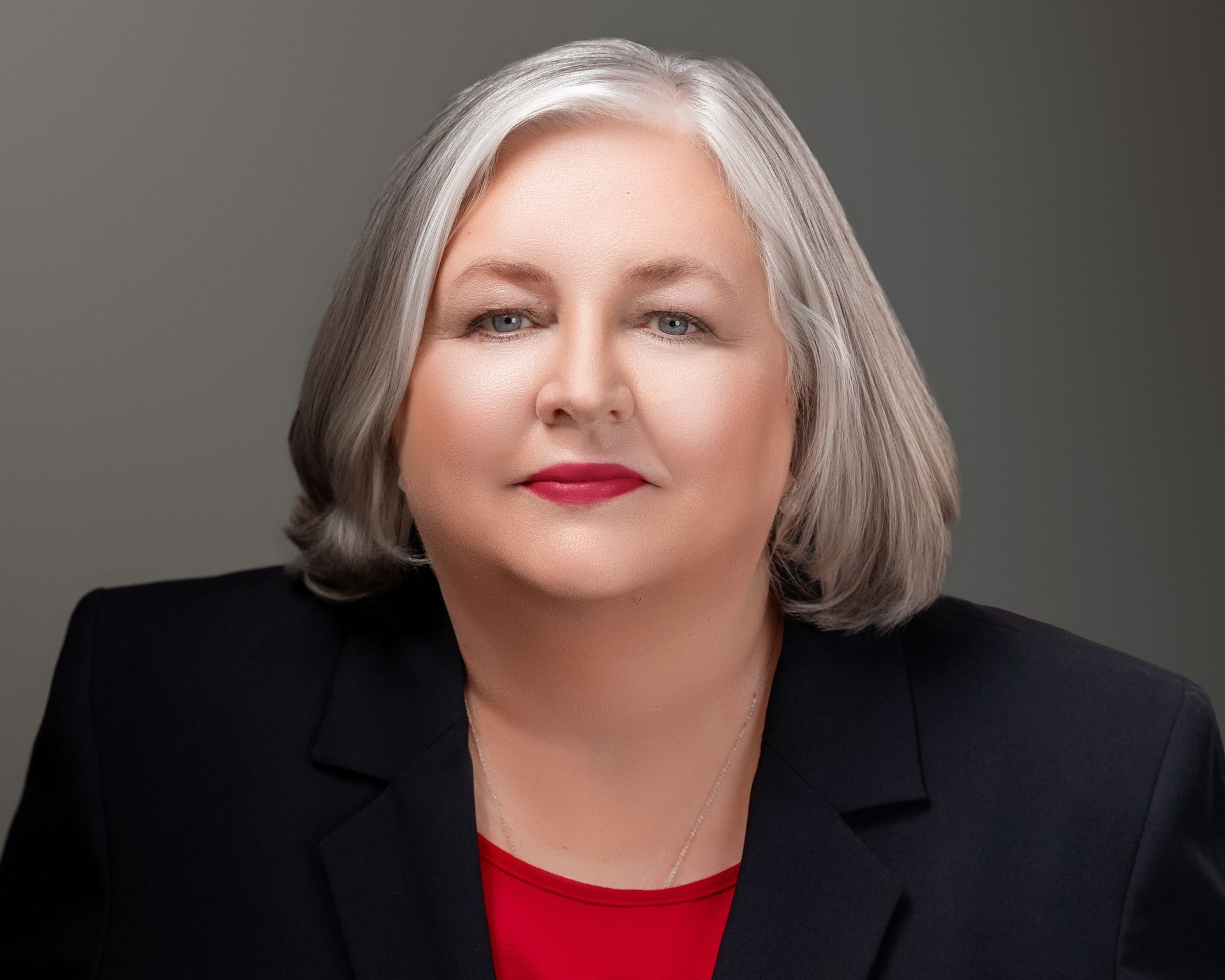 Portrait of a woman with gray hair in a black blazer and red top against a gray background