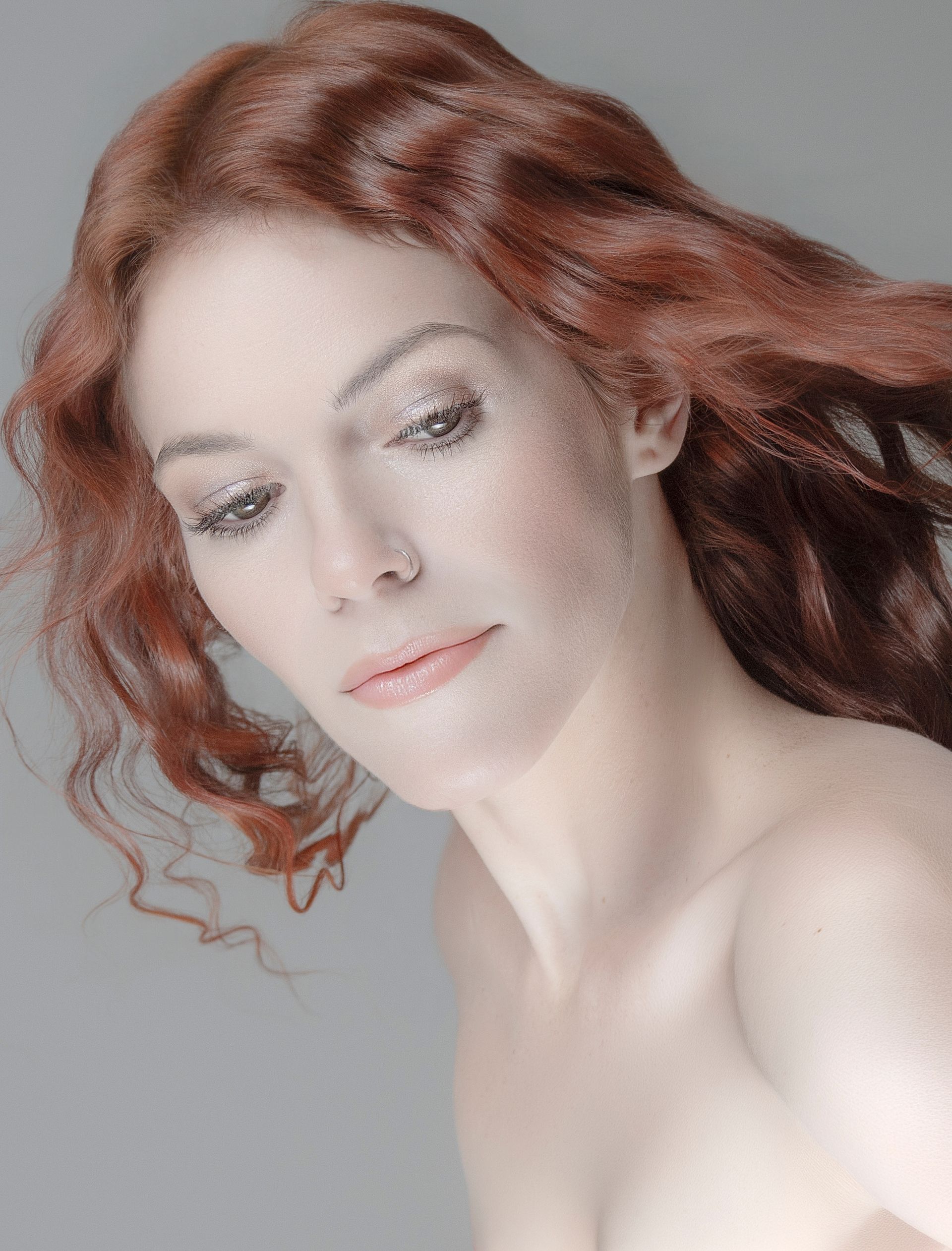 Portrait of a woman with wavy red hair against a gray background, looking down softly
