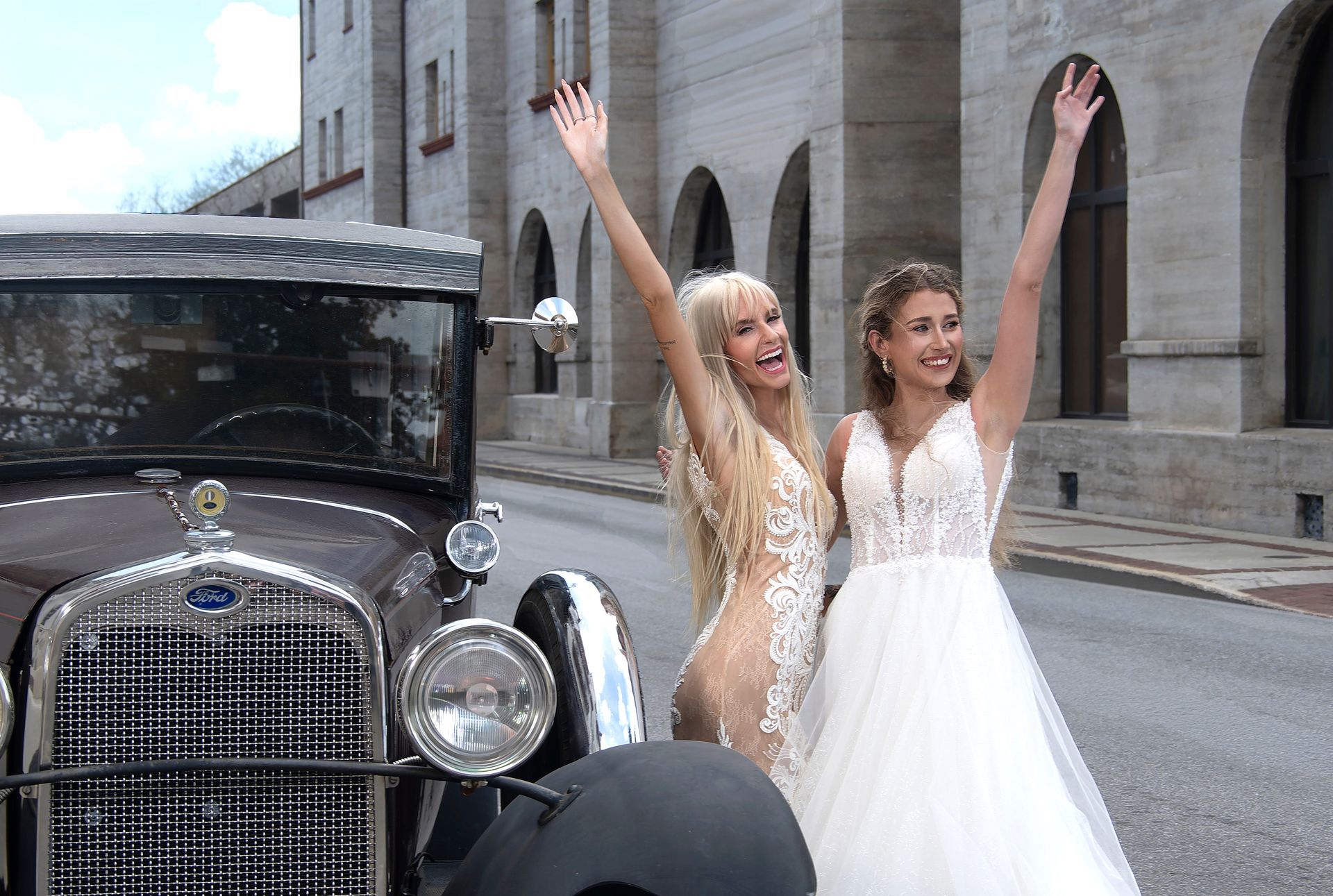 Two women in white dresses cheering beside a vintage black car on a city street